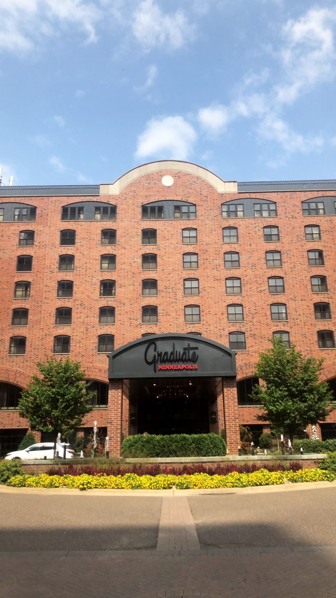 A large brick building with multiple windows stands under a blue sky, welcoming first-timers. The entrance features an arched canopy with "Graduate Minneapolis" written on it. Two trees and a flowerbed frame the entrance, offering a charming backdrop for WordCamp attendees.