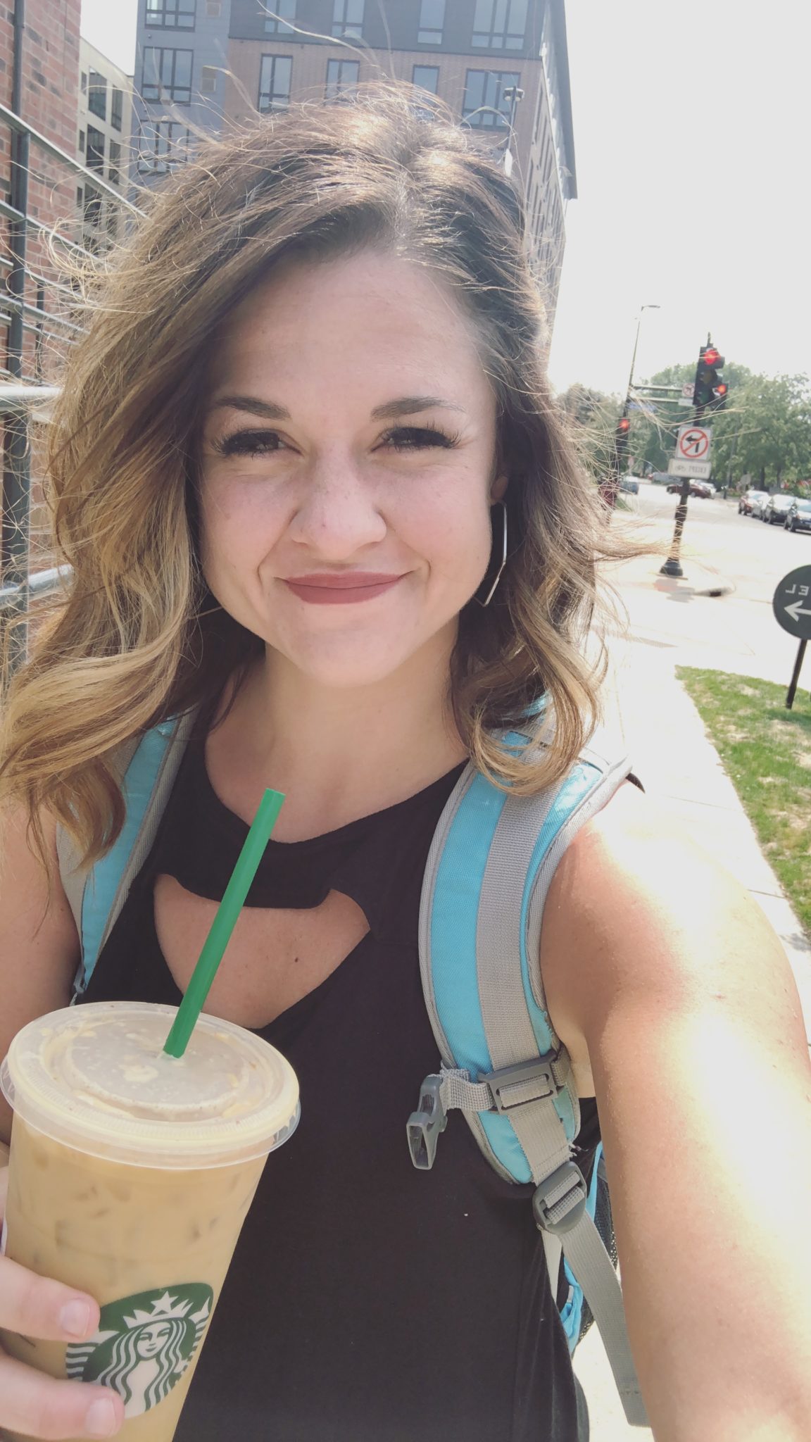 A woman with wavy hair smiles at the camera while holding a large iced coffee. As a WordCamp first-timer, she sports a black sleeveless top and backpack, standing outdoors on a sunny day with buildings and trees in the background.