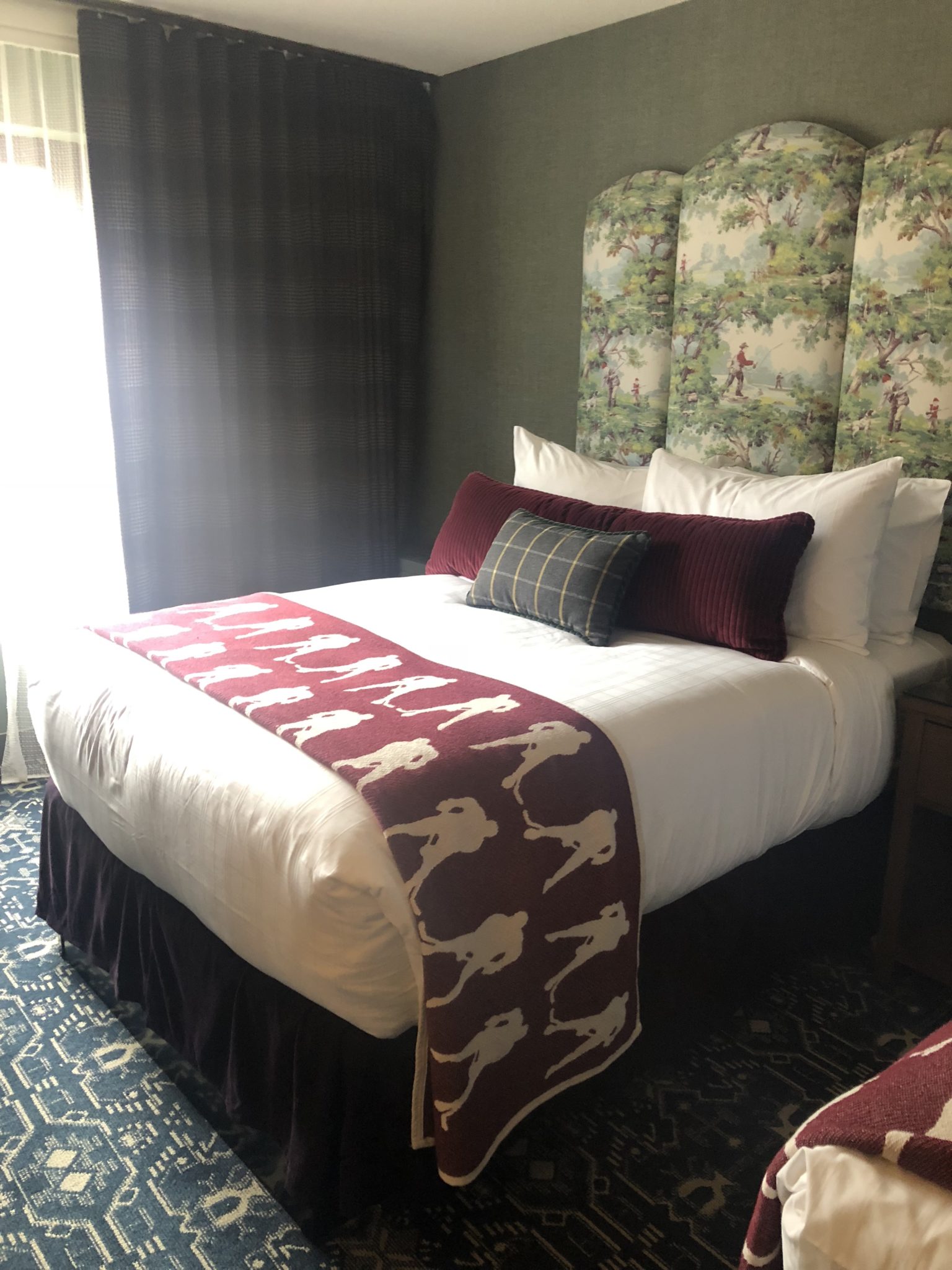 A cozy bedroom welcomes first-timers with a plush bed, white linens, and a maroon throw adorned with a horse pattern. The headboard flaunts a floral design, while dark curtains frame the window. A small decorative pillow sits atop the bed, complementing the patterned carpet.