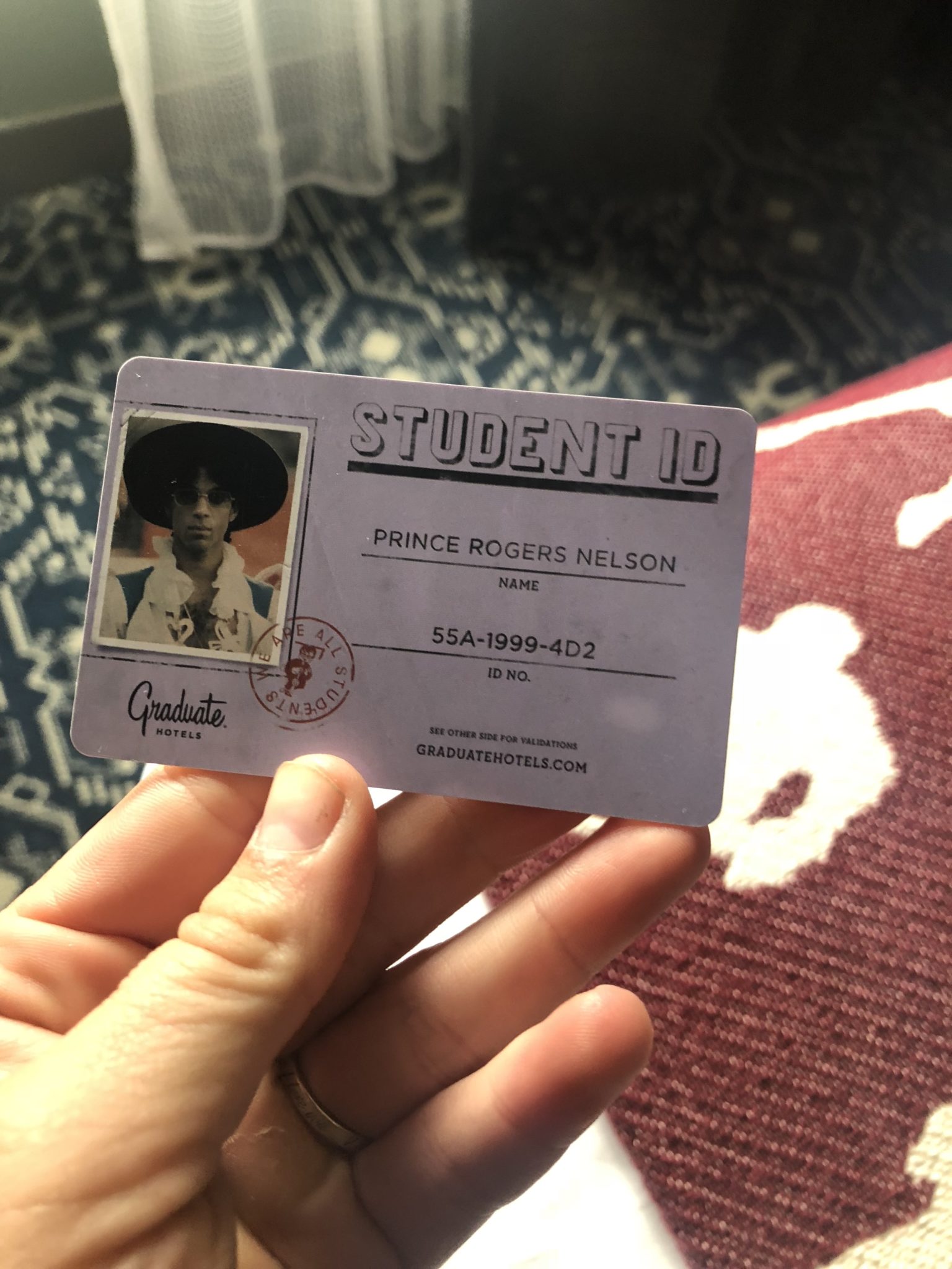 A first-timer holds a novelty student ID card with "Graduate Hotels" branding, featuring a photo labeled "Prince Rogers Nelson" and ID number "55A-1999-4D2." The card stylishly contrasts against a patterned fabric background, perfect for sharing at WordCamp.