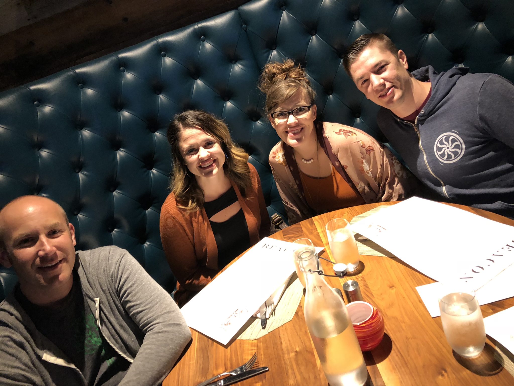 Four first-timers are seated at a round table with menus at a restaurant, smiling and facing the camera. The booth has a blue, tufted backrest. A bottle of water, glasses, and a small container are on the table, as they discuss their recent experiences from WordCamp.