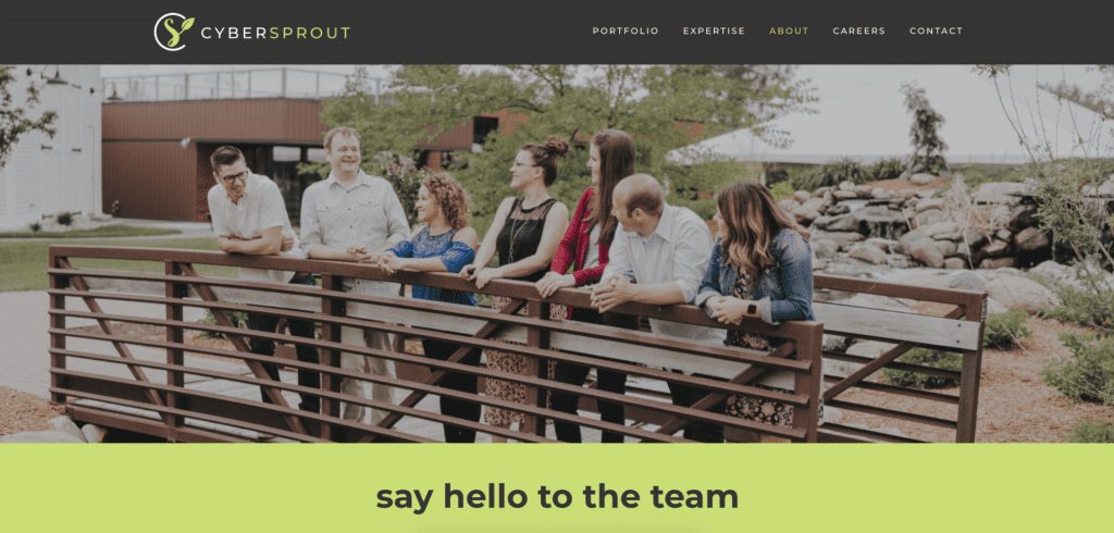 A group of seven people stand on a wooden bridge outdoors, smiling and engaged in conversation. The backdrop features greenery and a modern building, as if choosing the perfect site for inspiration. "CYBERSPROUT" headlines the scene, with menu options above "say hello to the team.