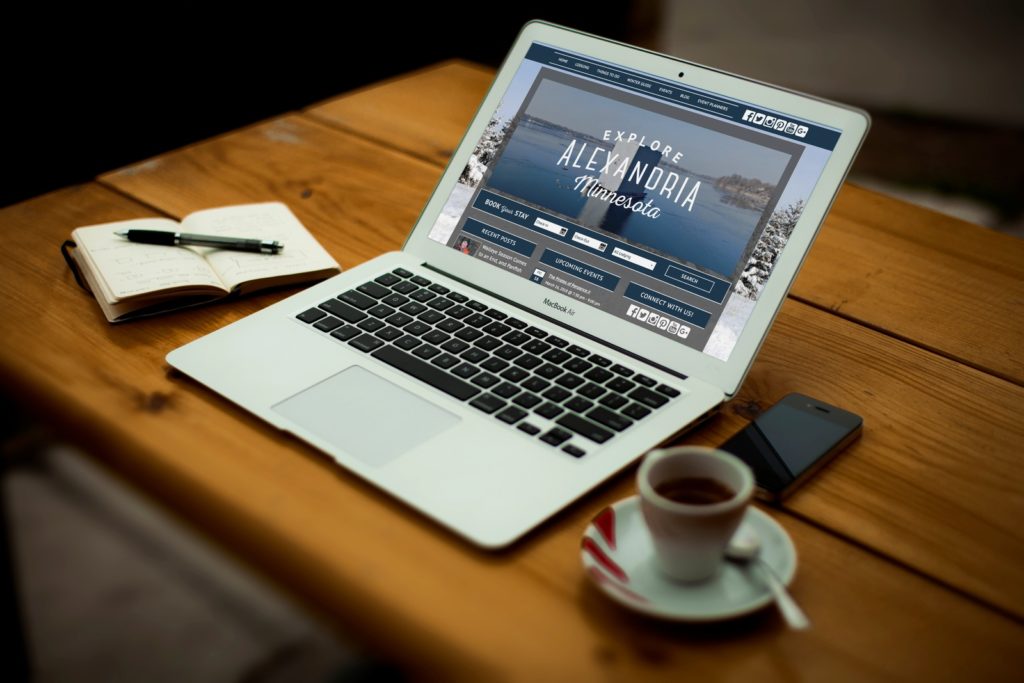 Displayed on a laptop, tablet, and smartphone, the website invites you to explore Alexandria, Minnesota. It features a scenic image underscoring its vibrant tourism potential with options for booking a stay and discovering local attractions.