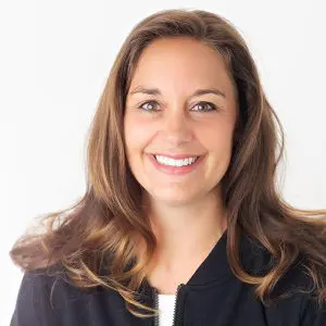 Brooke Erstad, a woman with long brown hair, smiles at the camera. She is wearing a black jacket over a white top, and the background is plain white.