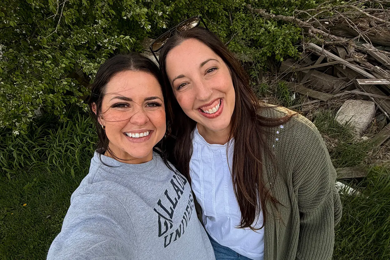 Two women smiling outdoors, standing close together for a selfie. One wears a gray Villanova University sweatshirt, the other a white shirt with a green sweater. Green grass and branches are visible in the background, giving the scene a fresh and lively feel reminiscent of posts from an old archive or snapshots taken outside a greenhouse.