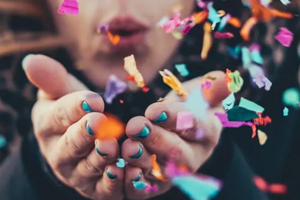 A person with teal nail polish blows colorful confetti from their cupped hands, creating a festive and vibrant scene like a celebration in the archive of joyful posts, with blurred confetti pieces floating through the air.