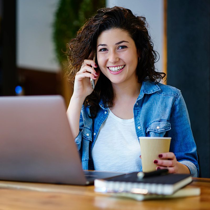 A smiling woman with curly hair is sitting at a table, holding a paper cup and talking on her smartphone. She is wearing a denim shirt over a white top, and an open laptop and a notebook are in front of her.