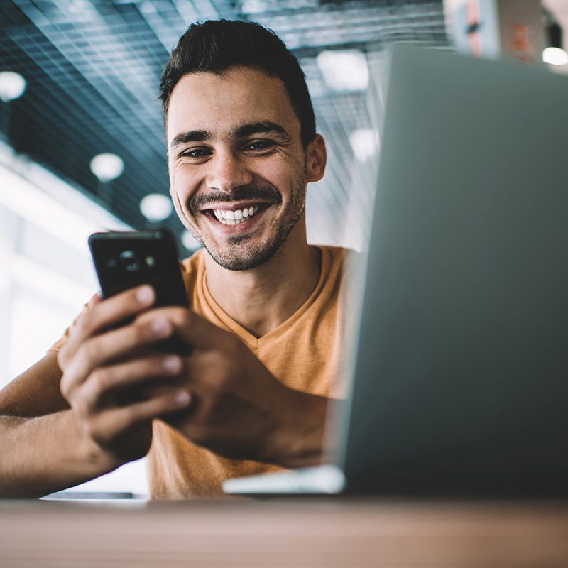A man with short dark hair and a beard is smiling while looking at his phone. He is sitting at a table with a laptop in front of him. The background features a modern interior with blurred lights.