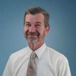 Smiling middle-aged man, Chris McConn, with short gray hair, wearing a cream-colored dress shirt and patterned beige tie, poses against a plain blue background.