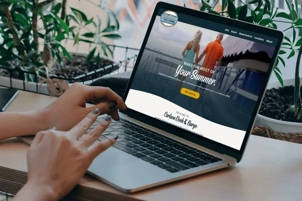 A person using a laptop at a wooden table with plants nearby; the screen displays a Carlson Dock website featuring two people on a dock with the text “Make the most of your summer!” and a yellow button.