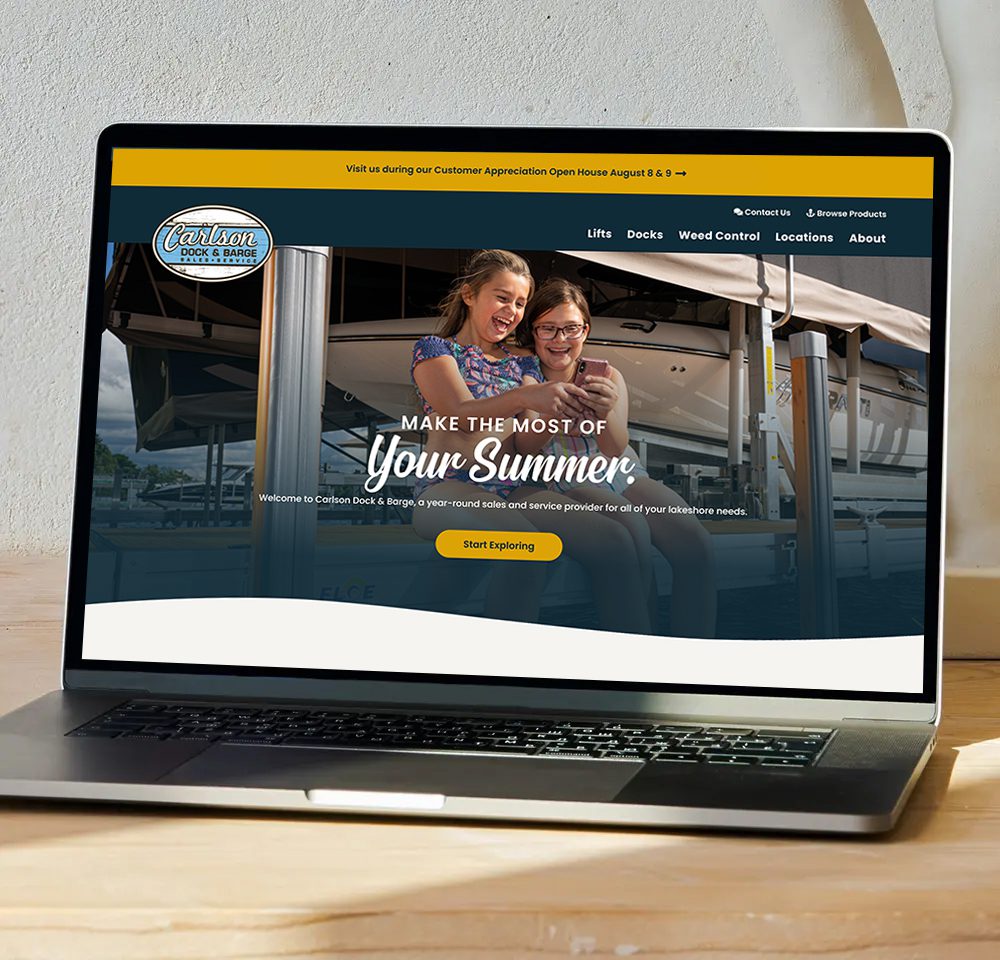 A laptop displays the Carlson Dock & Barge website, showing two smiling girls taking a selfie at a dock with boats in the background and the text "Make the Most of Your Summer" on the screen.