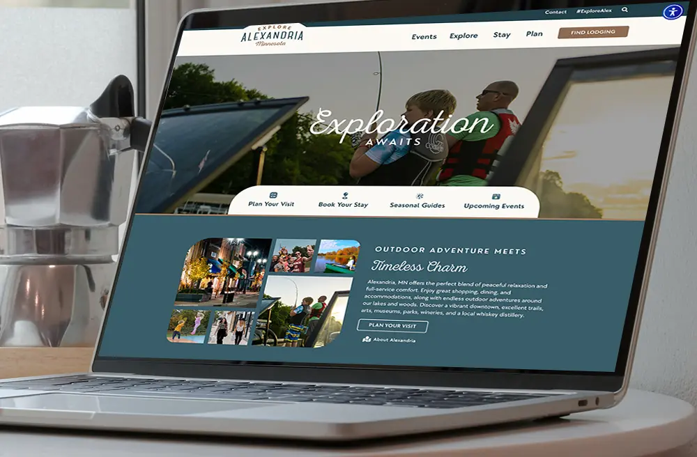 A laptop on a desk displays a travel website inviting users to explore Alexandria, Minnesota, with outdoor adventure images and the headline “Exploration Awaits.” A coffee maker sits beside the laptop.