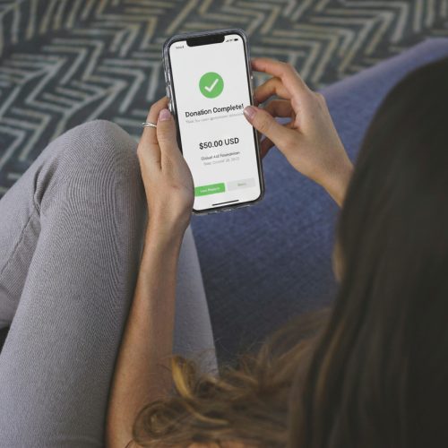 A person holding a smartphone displays a confirmation screen for a $50 USD donation, powered by WordPress donation tools for nonprofits, with a green checkmark and "Donation Complete!" while sitting cross-legged on a blue couch.