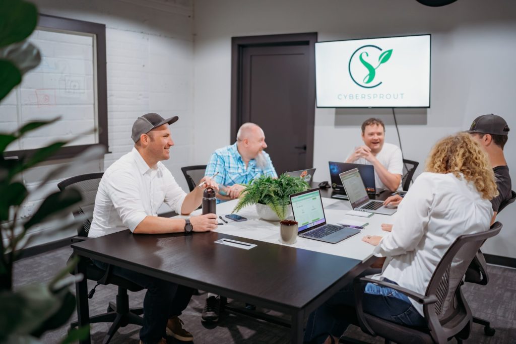 Five small business owners sit around a conference table with laptops, having a discussion. A screen on the wall displays the Cybersprout logo. The atmosphere appears casual and collaborative as they share tips to build your own website.