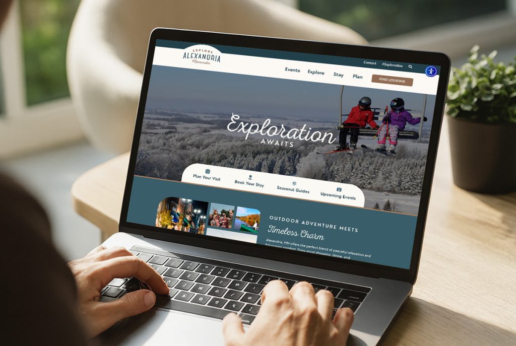 A person uses a laptop displaying a travel website featuring winter activities in Alexandria, with an image of two children playing in the snow and a headline that reads "Exploration Awaits.