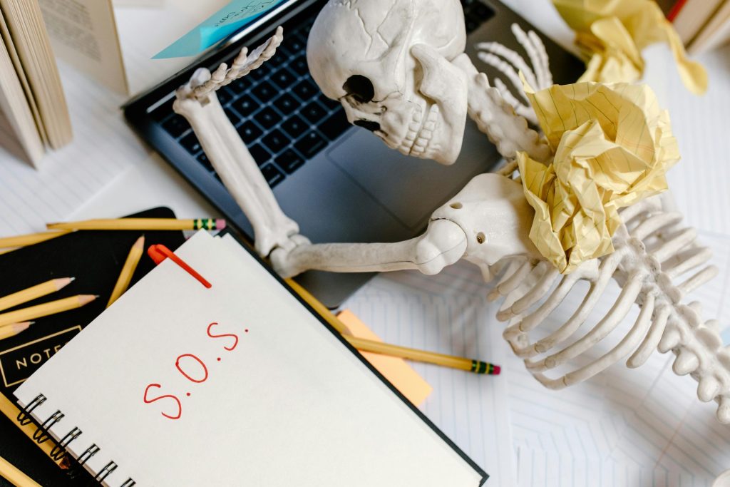 A plastic skeleton slumps over a laptop and messy desk, surrounded by crumpled papers and a notebook scrawled with "S.O.S."—perfectly capturing how small business owners feel when trying to build your own website.