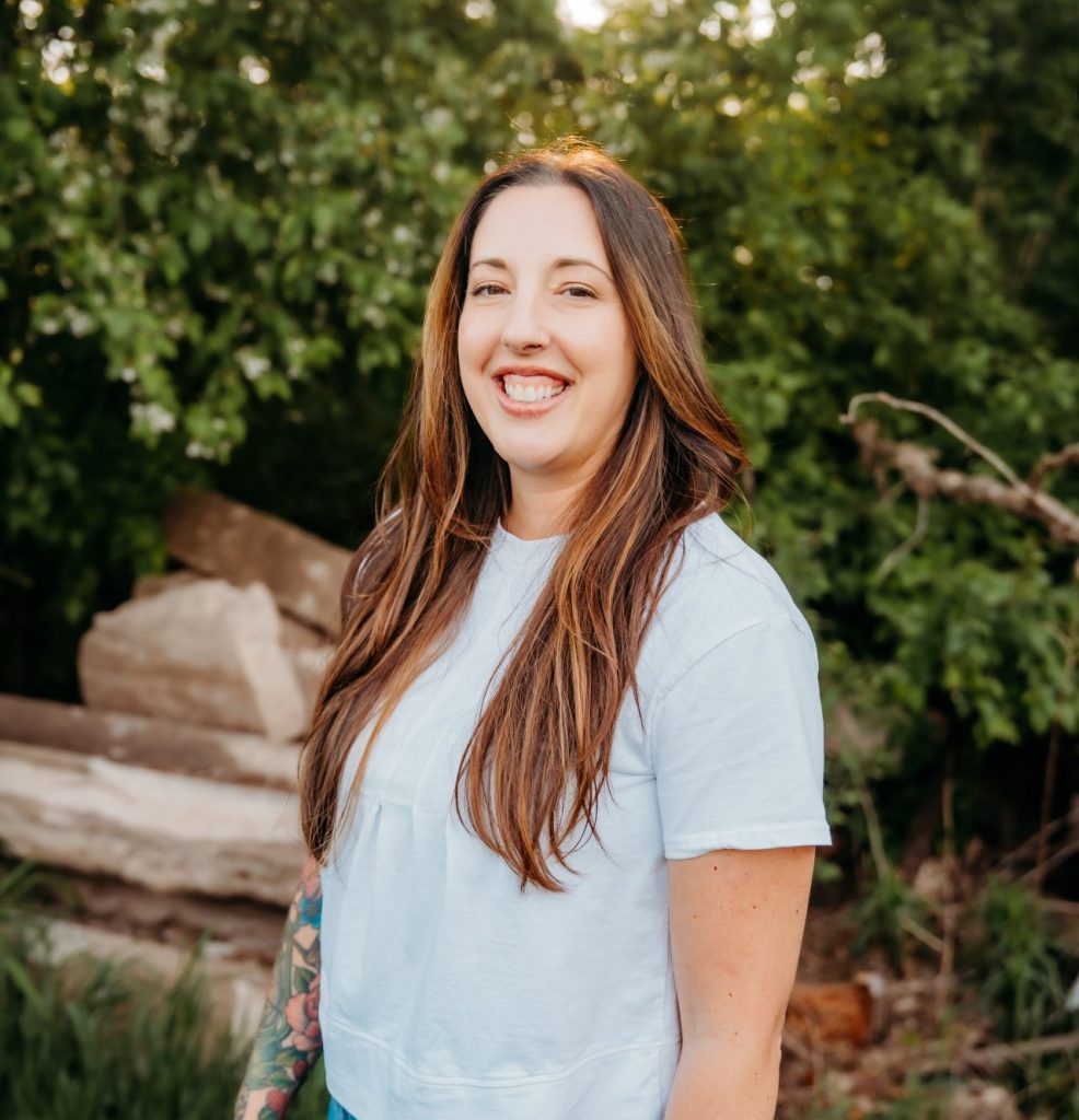 Tiffany Jenniges, with long brown hair, stands outside smiling in a light blue shirt and denim skirt. A tattoo decorates her left forearm as sunlight filters through green trees and rocks behind her.