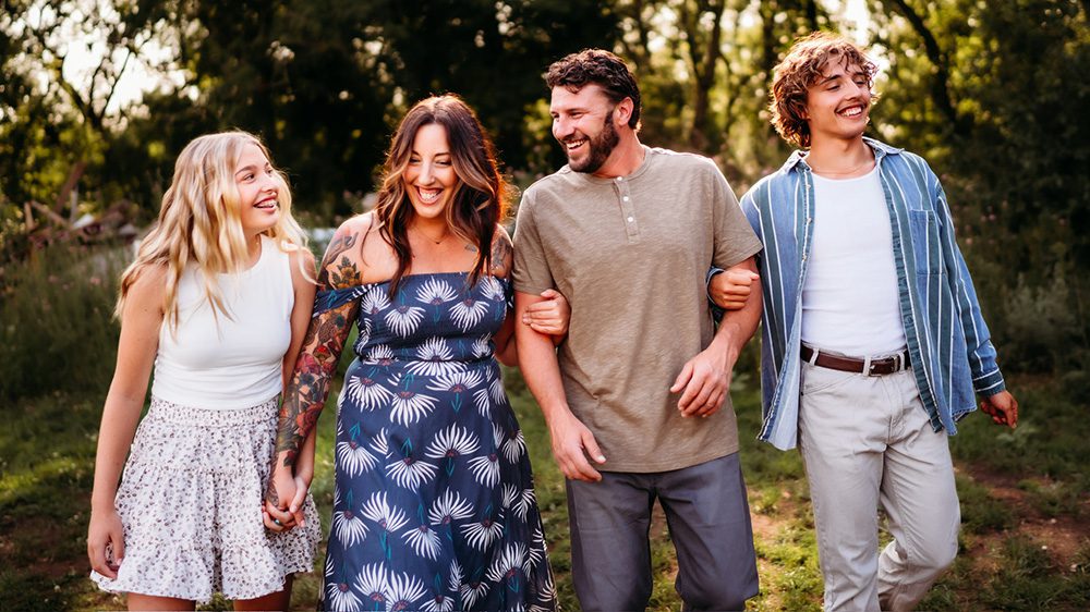 Four people walk arm in arm outdoors, smiling and laughing. They appear to be a family—perhaps even Tiffany Jenniges, Web Solutions Consultant at CYBERsprout, enjoying a sunny day together in a natural, green setting.