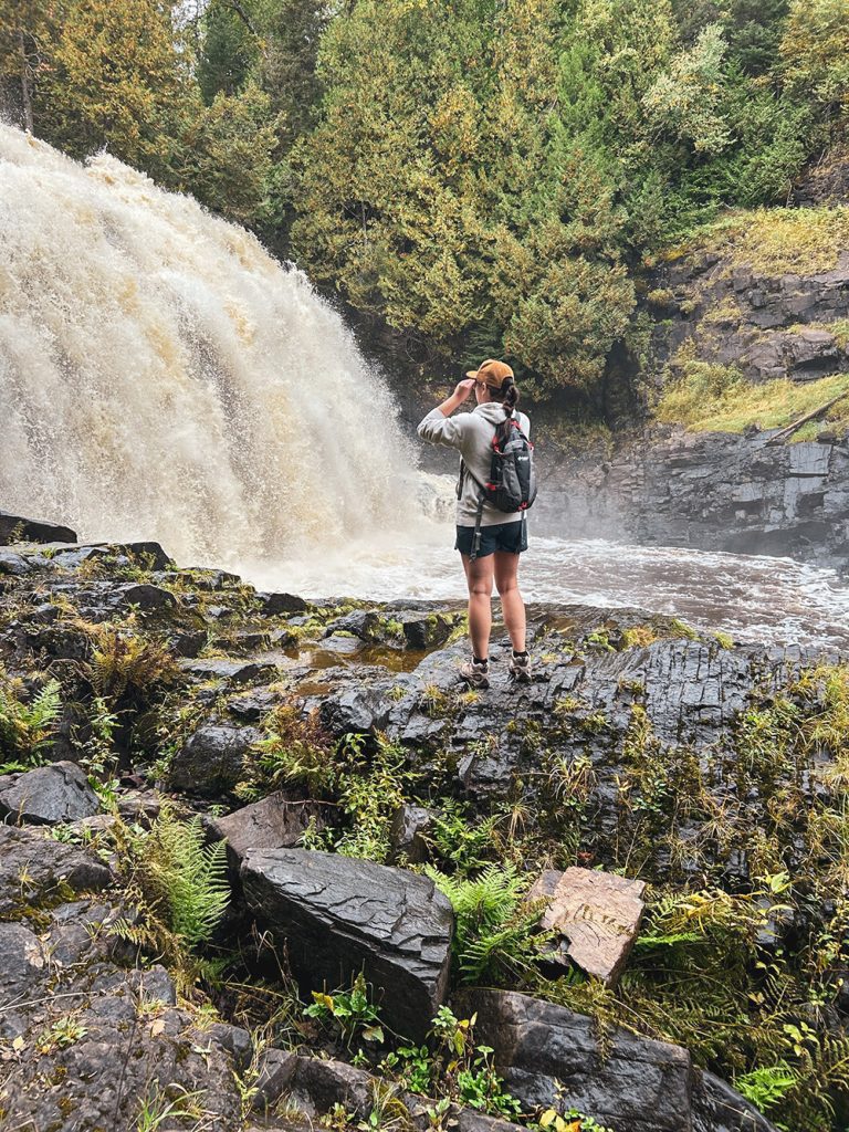 A person wearing a backpack stands on rocky terrain, looking at a large waterfall surrounded by trees and greenery—capturing the adventurous spirit that inspires Tiffany Jenniges, Web Solutions Consultant at CYBERsprout.