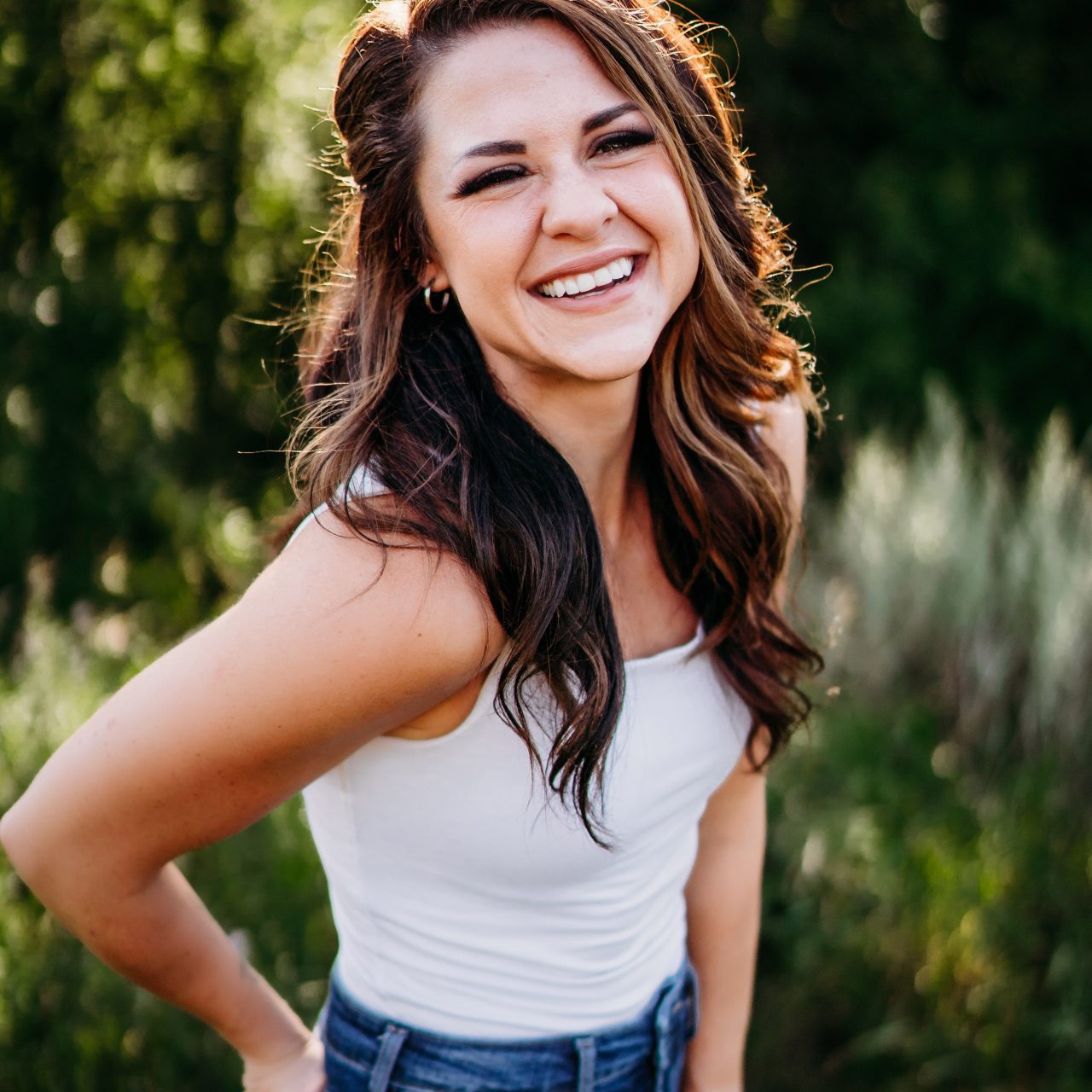 A woman with long brown hair is smiling and posing outdoors. She is wearing a white sleeveless top and blue jeans. The background features green foliage and sunlight filtering through the trees.